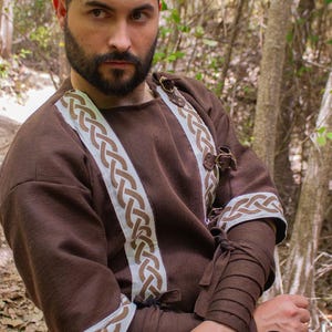 May include: A brown tunic with white and brown braided trim. The tunic has a round neckline and long sleeves. The sleeves have a brown fabric band at the wrist. The tunic is fastened with leather straps and buckles.