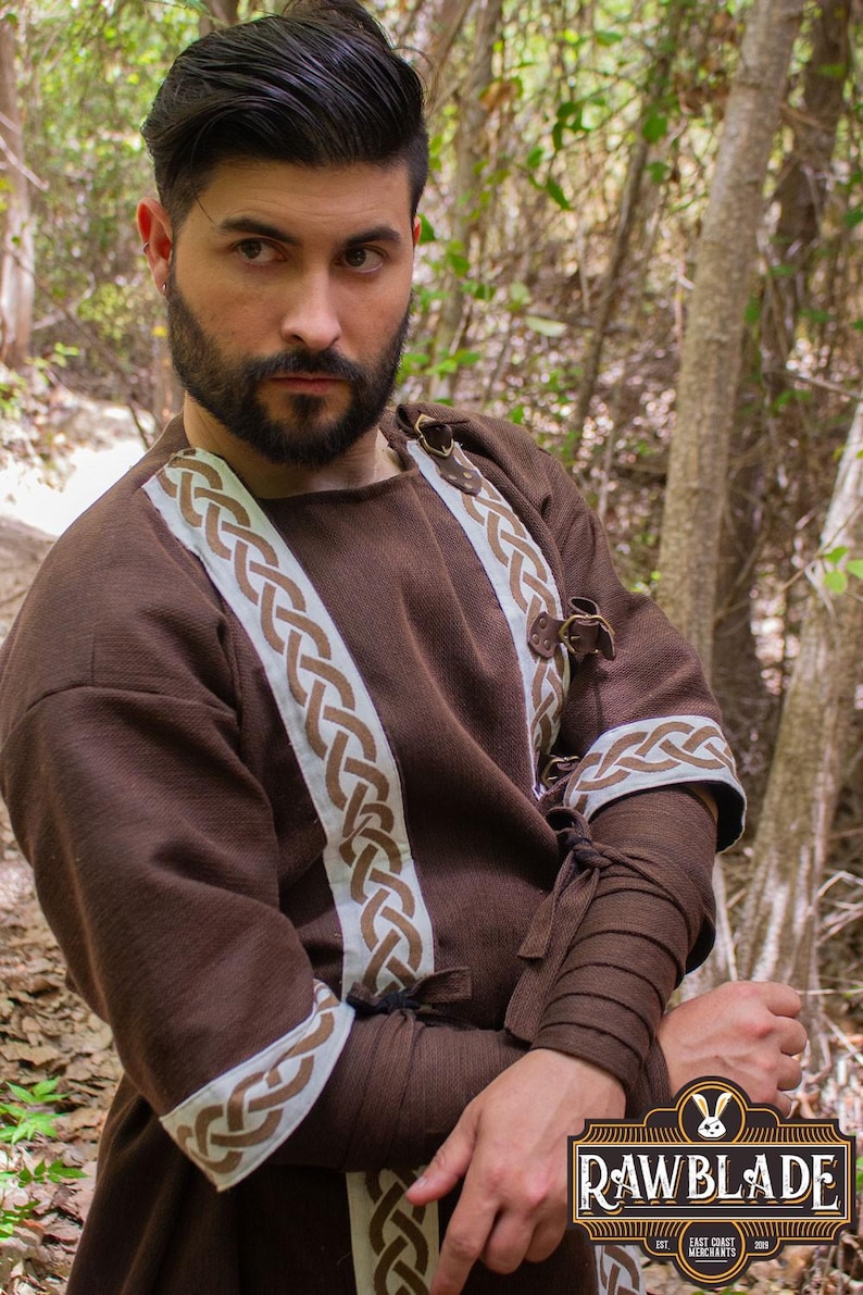 May include: A brown tunic with white and brown braided trim. The tunic has a round neckline and long sleeves. The sleeves have a brown fabric band at the wrist. The tunic is fastened with leather straps and buckles.