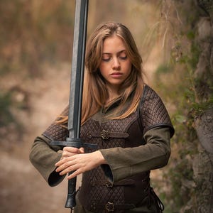 May include: A woman in a brown leather vest and green shirt holds a black sword. The sword has a long, straight blade and a black handle. The woman is standing in a forest setting.