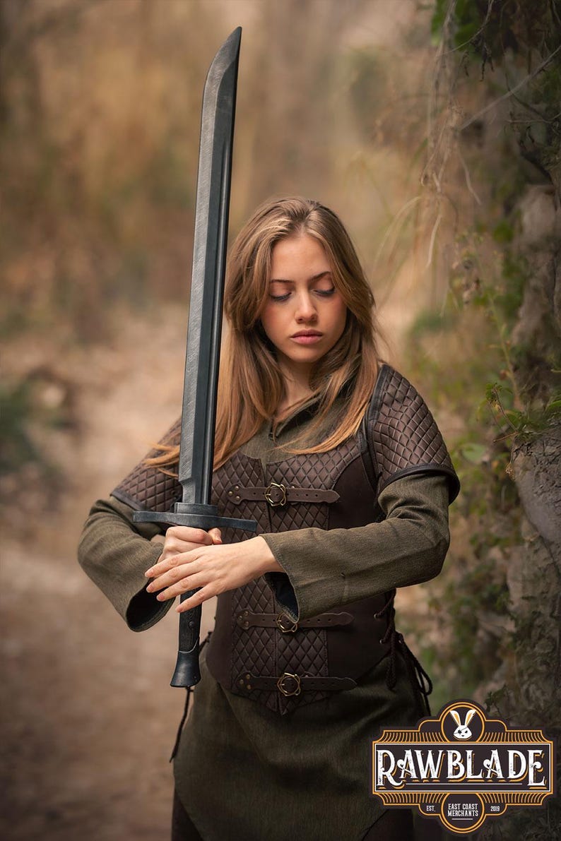 May include: A woman in a brown leather vest and green shirt holds a black sword. The sword has a long, straight blade and a black handle. The woman is standing in a forest setting.