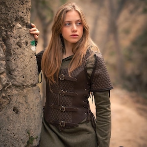May include: A brown leather vest with a quilted design and multiple buckles. The vest has a lace-up front and is worn over a green shirt.