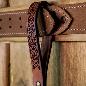 May include: A brown leather belt with a decorative, black, floral pattern. The belt has a metal buckle and is hanging on a wooden surface. The text "Rawblade East Coast Merchants Est. 2019" is visible in the lower right corner of the image.