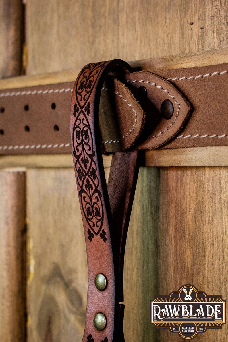 May include: A brown leather belt with a decorative, black, floral pattern. The belt has a metal buckle and is hanging on a wooden surface. The text "Rawblade East Coast Merchants Est. 2019" is visible in the lower right corner of the image.