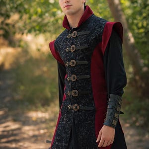 Peut inclure: Une tunique de style médiéval noire et rouge avec des détails ornés et des fermetures à boucle. La tunique présente un col haut et est portée sur une chemise noire à manches longues. Le logo Rawblade est visible en bas.