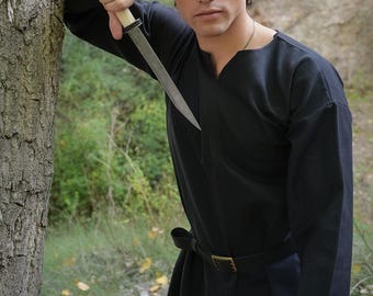 May include: A black tunic and a knife with a silver blade and a light-coloured handle. The tunic is belted at the waist. The logo "RAWBLADE" is visible at the bottom of the image. The person is leaning against a tree.