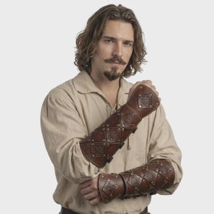 Pode incluir: Braçadeiras de couro castanho com padrões de losangos e quadrados, presas com rebites e fivelas. As braçadeiras de estilo medieval apresentam desenhos florais em relevo, oferecendo proteção e uma estética histórica. Estas braçadeiras são uma ótima adição a qualquer fantasia.