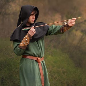 Puede incluir: Una persona que lleva una t&uacute;nica verde con una capa con capucha negra y un cintur&oacute;n de cuero marr&oacute;n, sosteniendo una flecha de madera con una pluma verde. La persona lleva brazaletes de cuero marr&oacute;n con detalles met&aacute;licos. El fondo es una imagen borrosa de follaje verde.