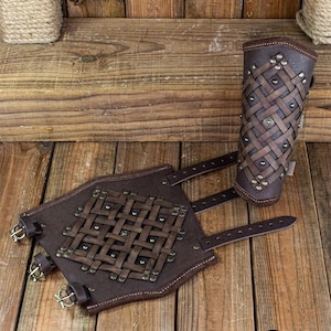 May include: A pair of brown leather arm guards with a woven design and metal accents. The guards are secured with straps and buckles.
