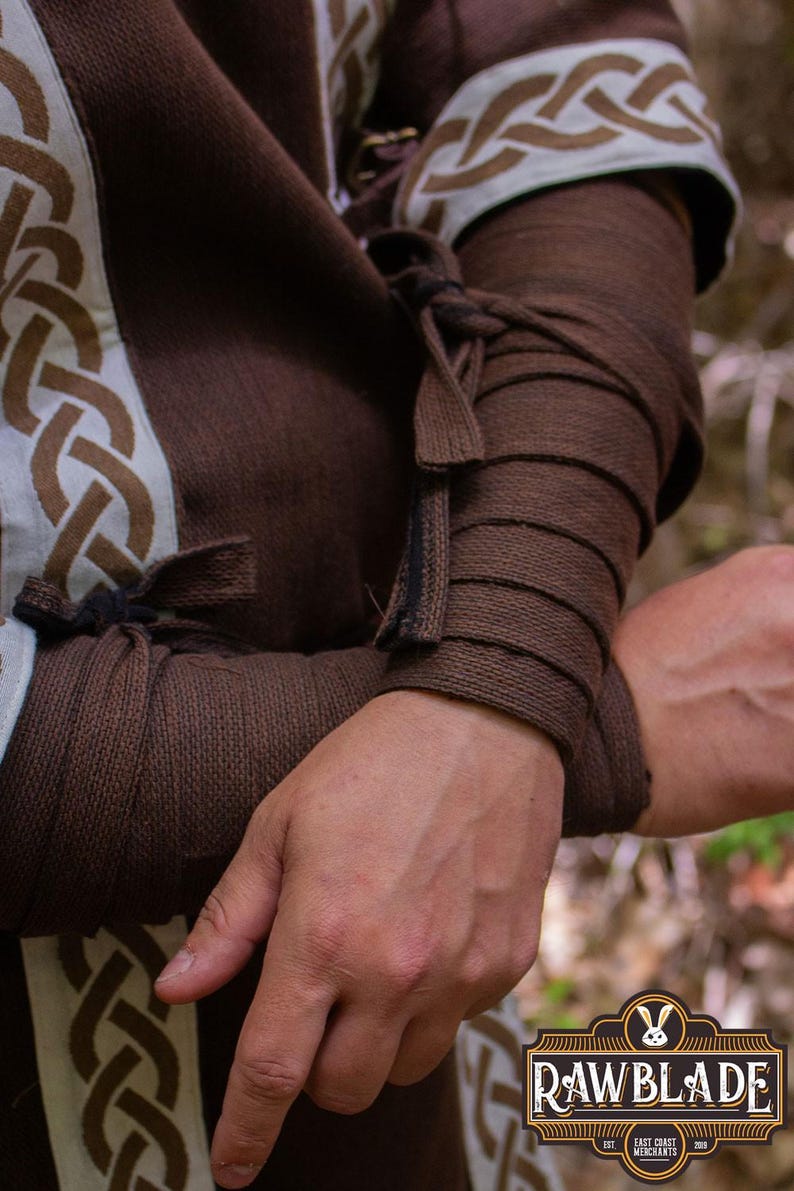 May include: Brown tunic with white and brown trim. The tunic has a Celtic knot design. The sleeves are rolled up and tied with brown fabric.