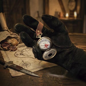 May include: A silver pocket watch compass with a red design is held by a gloved hand. The watch is open, revealing the compass face. A leather strap is attached to the watch. A map and a dagger are also visible.