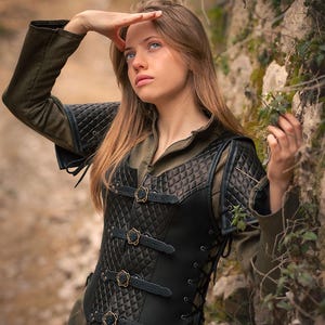 May include: A black leather corset with a quilted design and brass buckles. The corset has a lace-up back and is worn over a green shirt. The image is set against a backdrop of rocks and foliage.