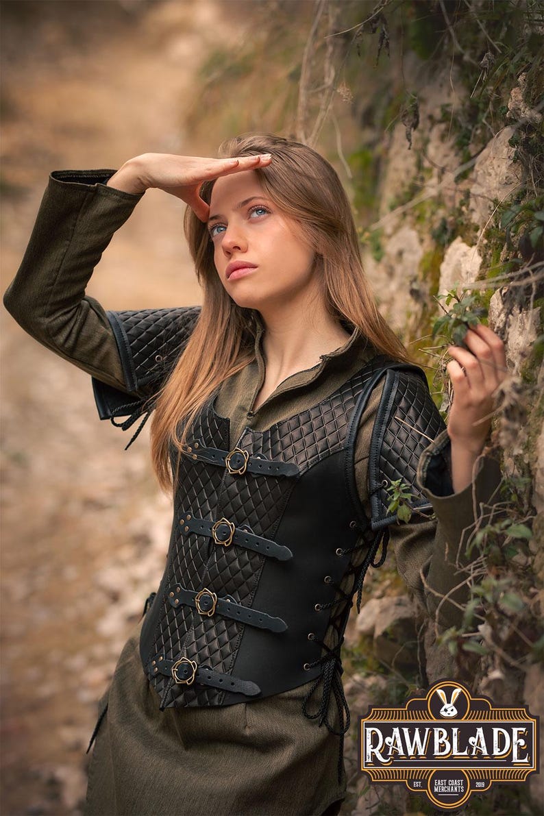 May include: A black leather corset with a quilted design and brass buckles. The corset has a lace-up back and is worn over a green shirt. The image is set against a backdrop of rocks and foliage.