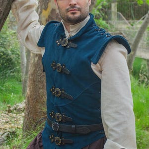 May include: A teal vest with black leather straps and brass buckles. The vest is worn over a cream-colored long-sleeved shirt and brown pants. The Rawblade logo is visible in the bottom right corner.