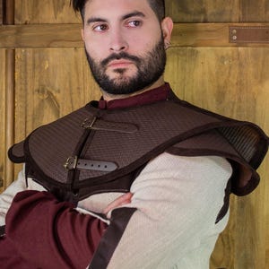 Könnte beinhalten: Eine braune Lederschulterrüstung mit Schnallen und Riemen. Die Rüstung wird über einem weißen und burgunderfarbenen Hemd getragen.