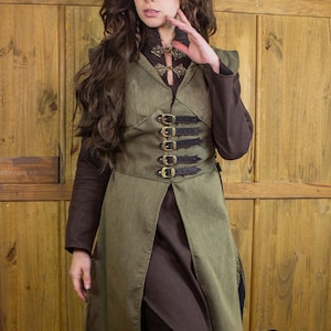 Pode incluir: Um colete comprido, verde, com capuz e uma roupa interior castanha. O colete tem uma fivela dourada decorativa e várias correias de couro preto. O colete é usado por uma pessoa com cabelo castanho comprido.