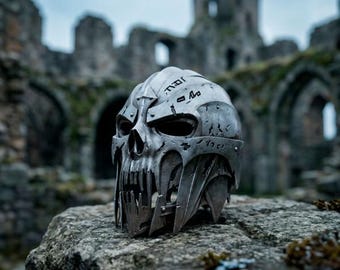 May include: A metallic silver skull-shaped helmet featuring jagged, tooth-like edges along the jawline, deep eye sockets, and engraved runic symbols across the forehead and sides. The surface has a weathered, distressed finish with visible rivets.