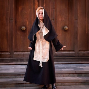 May include: A full-length black habit with a cream-coloured apron and headpiece. The garment features long sleeves, a high neckline, and a rope belt tied at the waist. A large, dark wooden Celtic cross pendant hangs from a cord around the neck.