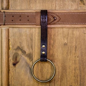 May include: A brown leather belt with a decorative metal ring attached. The belt is fastened to a wooden surface. The ring is made of brass and has a smooth finish.