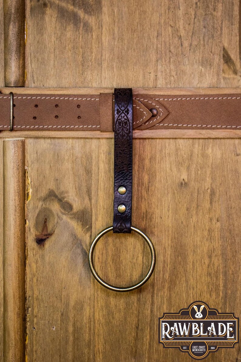 May include: A brown leather belt with a decorative metal ring attached. The belt is fastened to a wooden surface. The ring is made of brass and has a smooth finish.