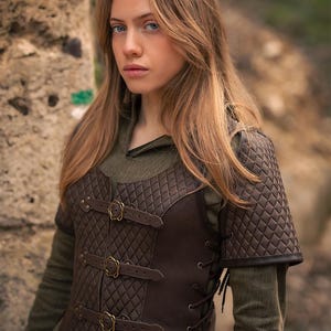 May include: A brown leather vest with a quilted pattern and multiple buckles. The vest has a lace-up back and is worn over a green shirt. The vest is labeled "Rawblade East Coast Merchants"