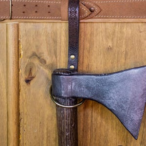 May include: A black axe with a wooden handle is mounted on a wooden wall with a brown leather strap. The axe has a metal ring around the handle. The strap has a decorative design and is attached to the wall with a metal buckle. The wall is made of wood planks. The axe is labeled "Rawblade East Coast Merchants Est. 2019."