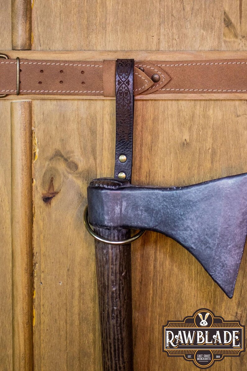 May include: A black axe with a wooden handle is mounted on a wooden wall with a brown leather strap. The axe has a metal ring around the handle. The strap has a decorative design and is attached to the wall with a metal buckle. The wall is made of wood planks. The axe is labeled "Rawblade East Coast Merchants Est. 2019."