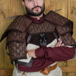 Könnte beinhalten: Braune Lederschulterrüstung mit Metallnieten und Riemen. Die Rüstung wird von einer Person mit Bart und einem dunkelroten Hemd getragen. Der Hintergrund ist eine Holzwand.