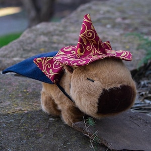 Puede incluir: Un capibara de peluche marrón que lleva un sombrero de mago rojo y dorado con una capa azul. El sombrero tiene un patrón de espiral dorado. El capibara está mirando hacia abajo. La imagen tiene el texto "Rawblade East Coast Merchants" en la parte inferior.