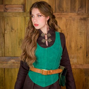 Puede incluir: Una camisa de manga larga marrón oscuro con cuello alto y dos cierres metálicos ornamentados, superpuesta con un chaleco de ante verde vibrante. El chaleco se ajusta a la cintura con un cinturón de cuero marrón claro con hebilla dorada.