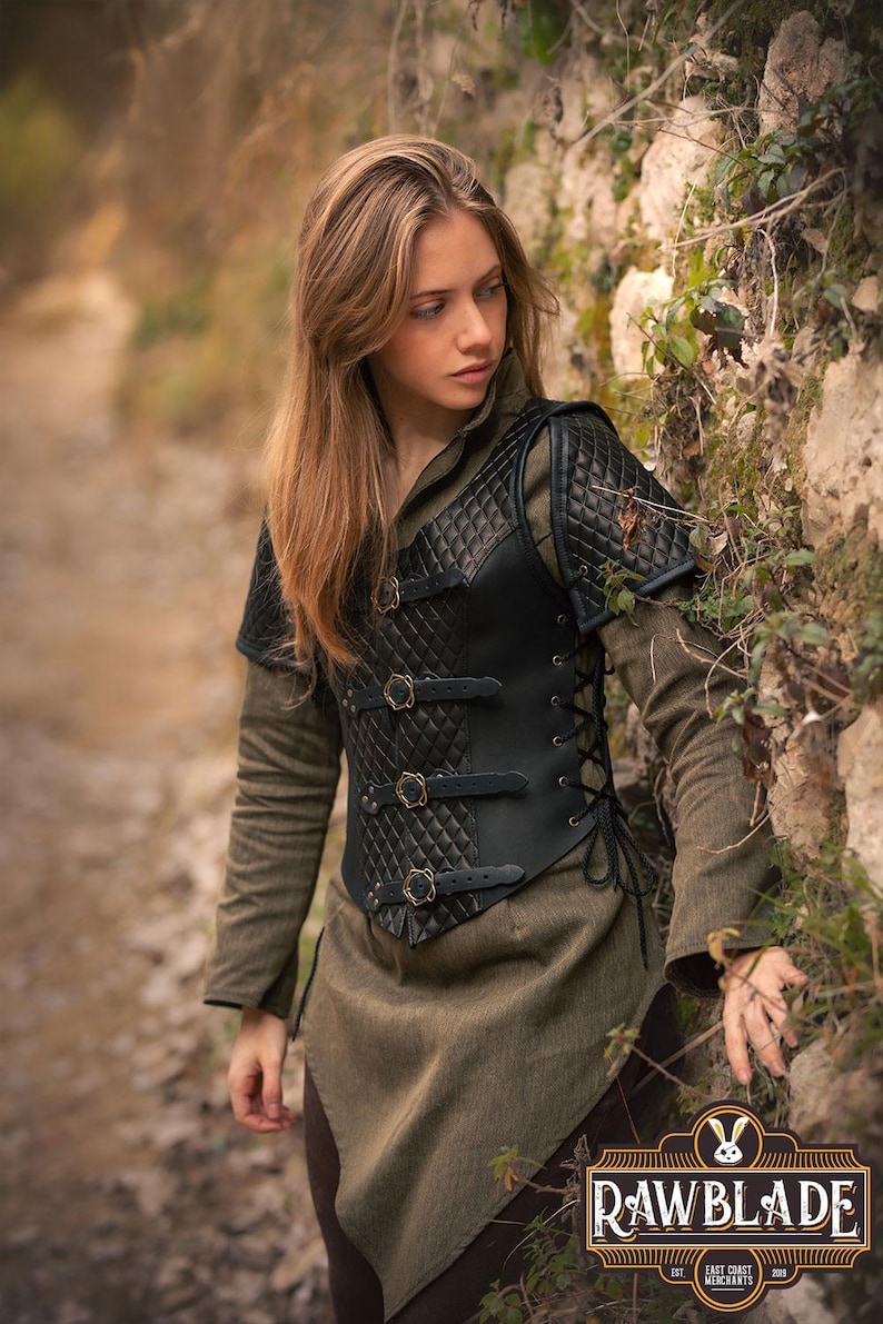 May include: A black leather quilted vest with a laced back and gold buckles. The vest has shoulder pads and is being worn by a person standing in a woodland setting.
