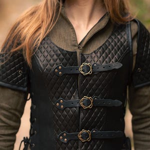 May include: Black leather vest with a quilted diamond pattern and gold buckles. The vest has a lace-up back and is worn over a green shirt. The vest is labelled "Rawblade East Coast Merchants 2018".