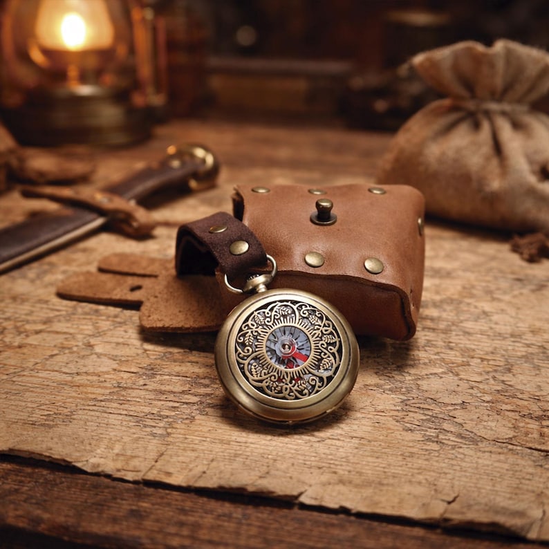 May include: A vintage-style pocket watch with an ornate, bronze-colored case and a detailed face. The watch is paired with a brown leather pouch and a matching strap. The watch is resting on a wooden surface, suggesting a steampunk or antique theme.