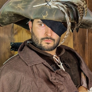 May include: A pirate hat with a wide brim, adorned with feathers, and a black eye patch. The hat is brown with a distressed finish. The person is wearing a brown cloak. The Rawblade logo is visible.