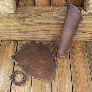 May include: Two brown leather arm guards with laces and eyelets. One arm guard is a full length sleeve and the other is a shorter cuff. A length of brown cord is included. The image includes the text "RAWBLADE EAST COAST MERCHANTS EST. 2018"