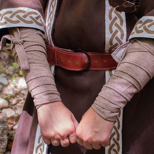 May include: A brown tunic with white and brown braided trim. The tunic has a brown leather belt with a metal buckle. The sleeves are rolled up and tied with a brown fabric. The person is wearing brown fabric arm wraps.
