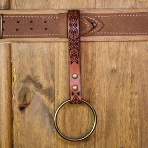 May include: A brown leather belt with a decorative metal buckle and a leather loop with a brass ring attached. The loop has an intricate floral design.
