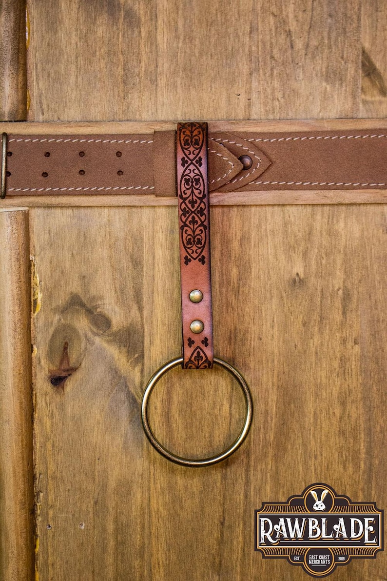 May include: A brown leather belt with a decorative metal buckle and a leather loop with a brass ring attached. The loop has an intricate floral design.