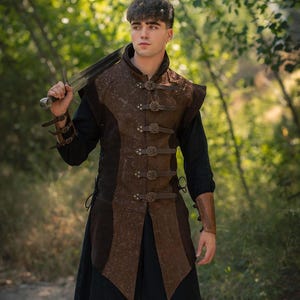 May include: A medieval-style brown leather vest with decorative buckles and a matching leather arm cuff. The vest is worn over a black long-sleeve shirt and black pants. A sword is held over the shoulder. The Rawblade logo is visible.