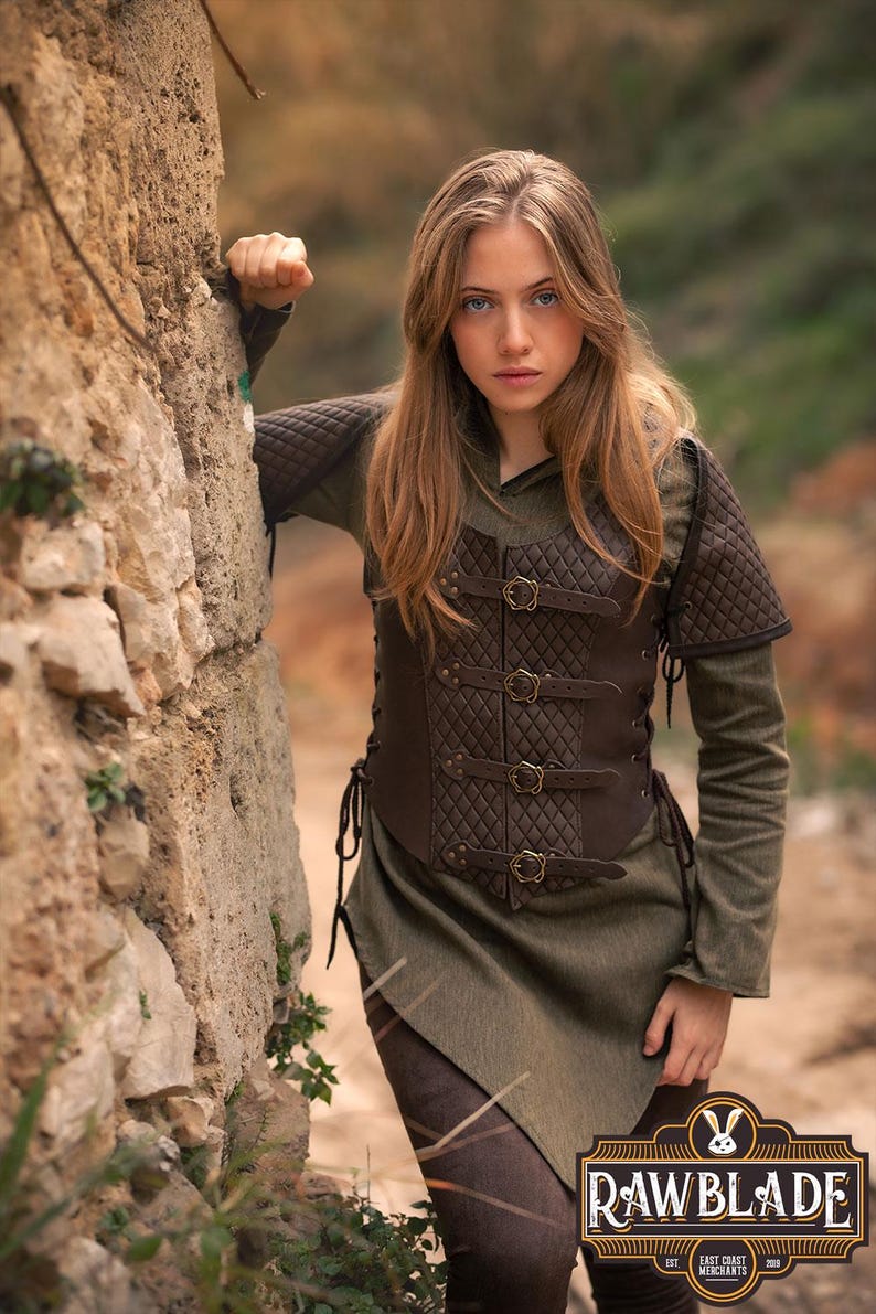 May include: A brown leather vest with quilted panels and buckles. The vest has a laced-up back and is worn over a green tunic. The model is standing next to a stone wall.