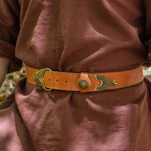 Pode incluir: Um cinto de couro castanho com uma fivela de latão que apresenta um design de árvore da vida. A fivela é ornamentada e tem um padrão de nó celta. O cinto é usado na cintura de uma pessoa que usa uma camisa castanha.