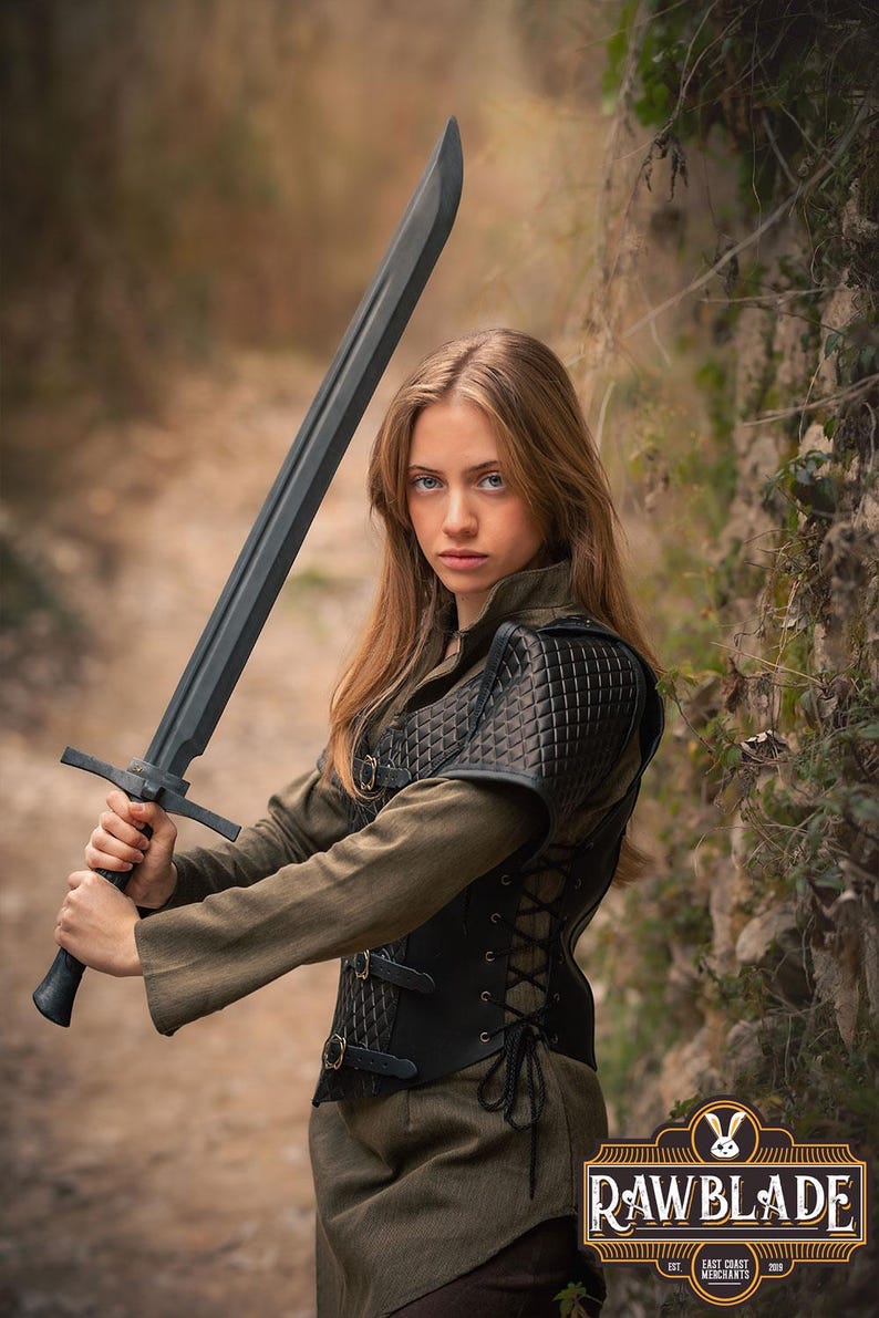 May include: A woman in a woodland setting holds a black sword with a leather-bound handle. She is wearing a black leather vest with a quilted design and a brown tunic. The vest has a lace-up back and buckles on the front. The background is a blurred image of trees and foliage.