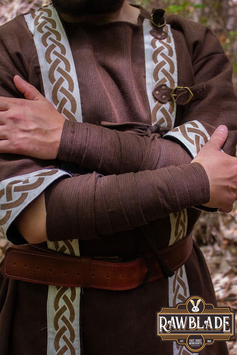 May include: A brown tunic with white and brown braided trim. The tunic has a brown leather belt with a buckle. The sleeves are rolled up and secured with brown fabric ties. The tunic is made of a soft, comfortable fabric.