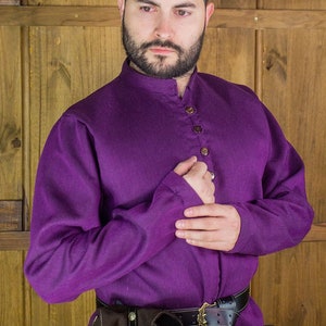 Könnte beinhalten: Ein langärmeliges, violettes Hemd mit Stehkragen und Knopfleiste. Das Hemd wird mit einem braunen Ledergürtel und einer braunen Ledertasche getragen.