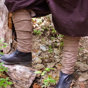 May include: A pair of brown fabric leg warmers worn over black leather boots. The leg warmers are folded over at the top and bottom. The person is wearing a dark purple skirt or pants. The image is taken in a forest setting.