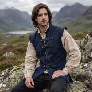 May include: A navy blue suede vest with black leather straps and buckles. The vest is worn over a cream-colored long-sleeved shirt. The background features a mountainous landscape under an overcast sky. The vest has a classic, historical design.
