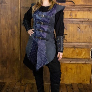 May include: A dark gray and blue quilted leather vest with a pointed hem and multiple buckles. The vest has a black lining and is worn over a black long-sleeved shirt.