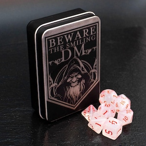 May include: A black metal box with a warning sign that reads "Beware the Smiling DM". The box is open and contains a set of pink and white dice with red numbers. The dice are translucent and have a cloudy appearance.