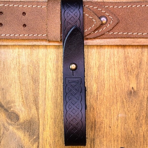 May include: A black leather belt loop with a Celtic knot design. The loop is attached to a brown leather belt with white stitching.