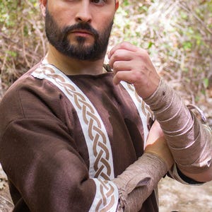 May include: A brown tunic with white and brown braided trim. The tunic has long sleeves and a round neckline. The model is wearing a brown leather belt.
