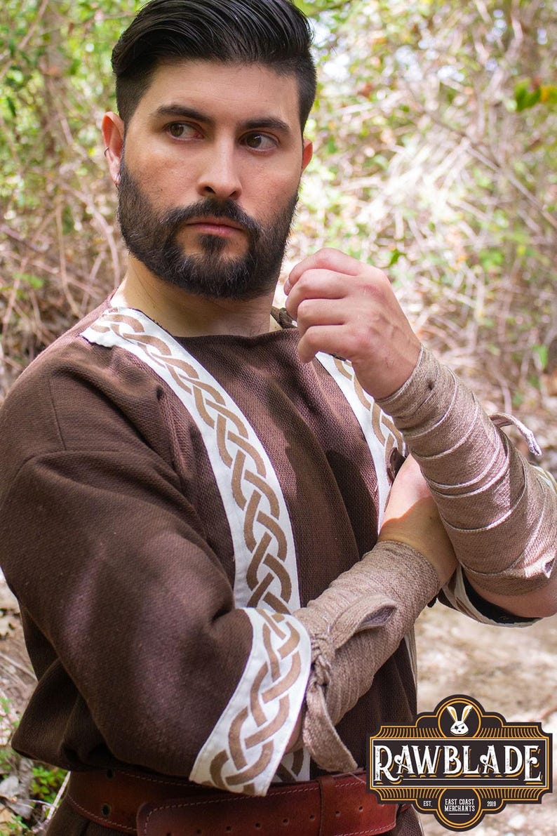 May include: A brown tunic with white and brown braided trim. The tunic has long sleeves and a round neckline. The model is wearing a brown leather belt.
